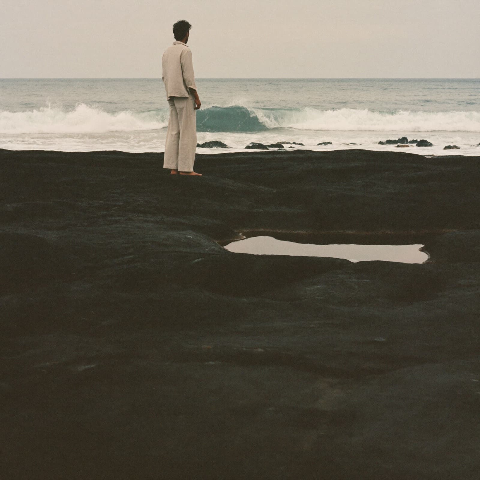Un homme seul, de dos, debout sur le basalte noir du littoral atlantique de Casablanca face à la houle, lumière d’un matin voilé